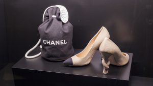 Valokuvassa jalustallaan Chanel-logolla varustettu musta merimieskassi sekä korkokengät, joiden korkoa koristaa solmun muoto.