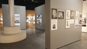 Valokuvassa on näyttelytila, jossa harmaita, valkoisia sekä puunvärisiä seiniä joille on ripustettu mustavalkoisia valokuvia.
