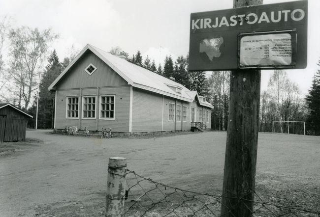 Taustalla puinen koulurakennus, iso tyhjä piha ja etualalla kirjastoautokyltti.